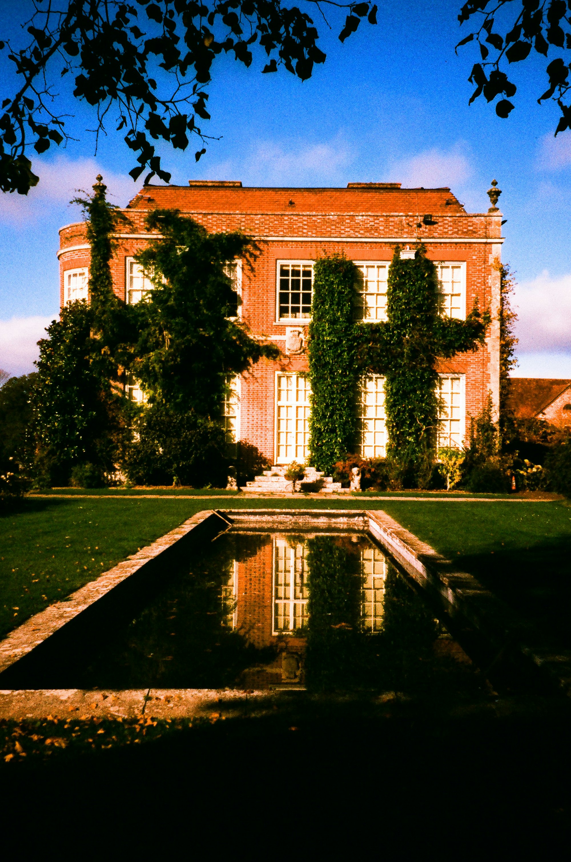 A red-brick manor house reflected in a long pool at golden hour